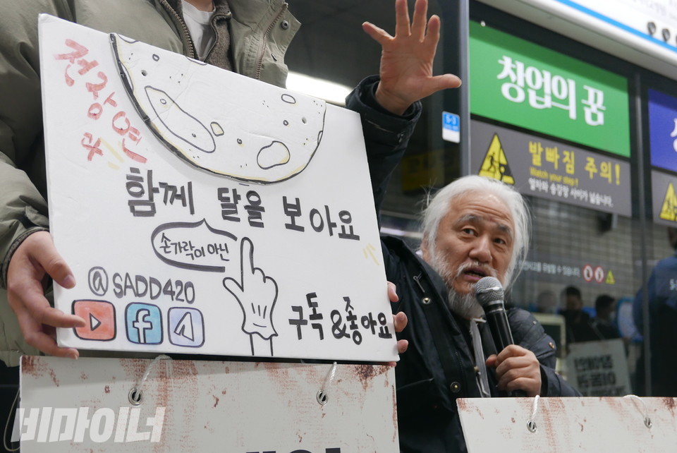 피켓에 달, 손 그림이 그려져 있다. 또한 “손가락이 아닌 전장연과 함께 달을 보아요. 구독 좋아요”라는 문구가 적혀 있다. 박경석 전장연 상임공동대표가 이와 관련된 얘길 하고 있다. 사진 양유진