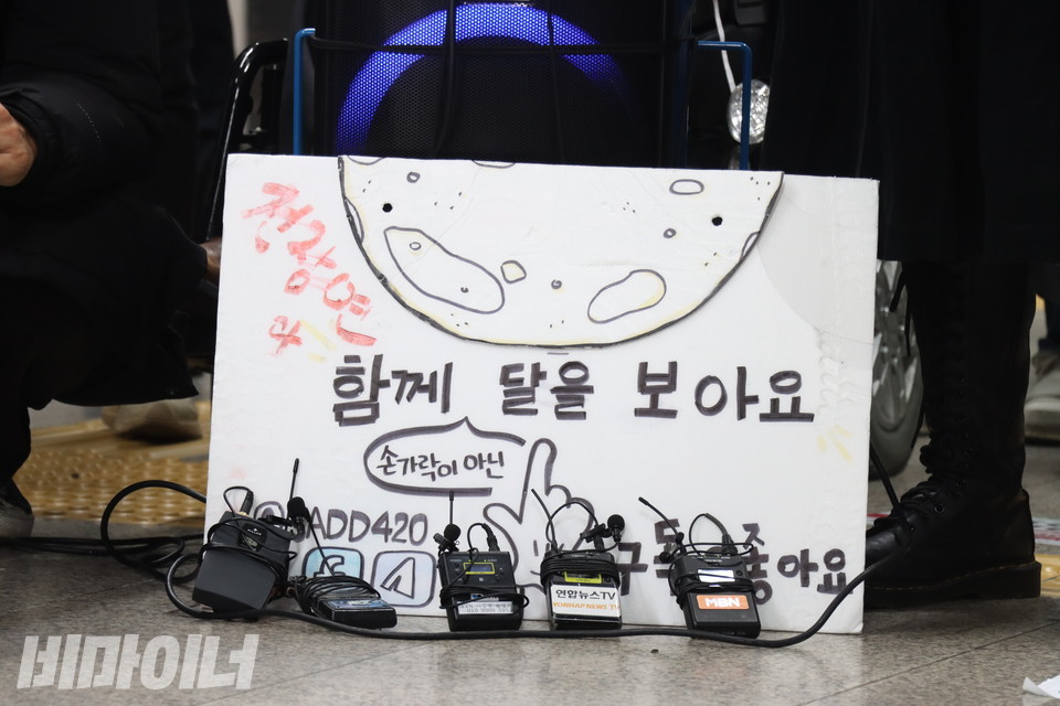 ‘전장연과 함께 손가락이 아닌 달을 보아요’라고 적힌 하얀 종이 앞에 방송국 마이크들이 올망졸망 기대어 있다. 사진 강혜민 
