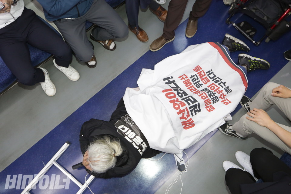 박경석 대표가 지하철에서 오체투지를 하고 있다. 그가 입고 있는 옷 뒷면에는 “이것도 노동이다, 권리중심중증장애인공공일자리”라고 적혀 있다. 그가 두르고 있는 흰 천에는 “예산 없이 권리 없다!”라고 적혀 있다. 사진 비마이너DB