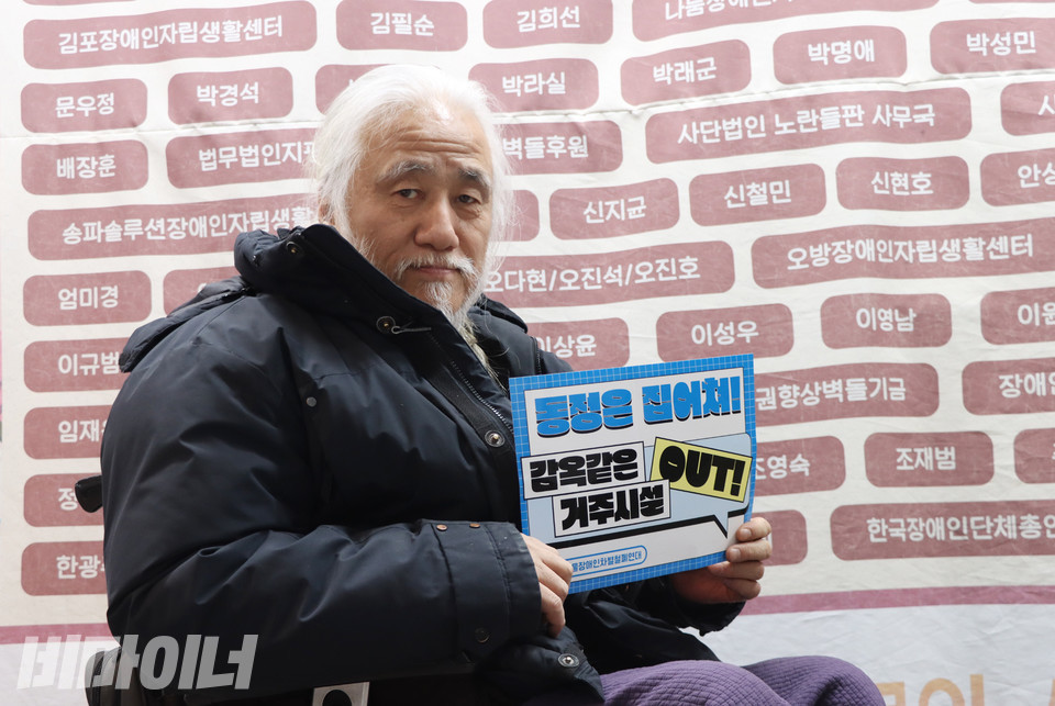 지난 18일, 박경석 대표가 인터뷰를 마친 후 “동정은 집어치워, 감옥 같은 거주시설 아웃(OUT)”이라고 적힌 피켓을 들고 있다. 그의 뒤에는 전장연 사무실이 대학로로 이사 올 때 임대료를 후원해준 ‘벽돌 회원’의 이름이 걸려 있다. 사진 강혜민