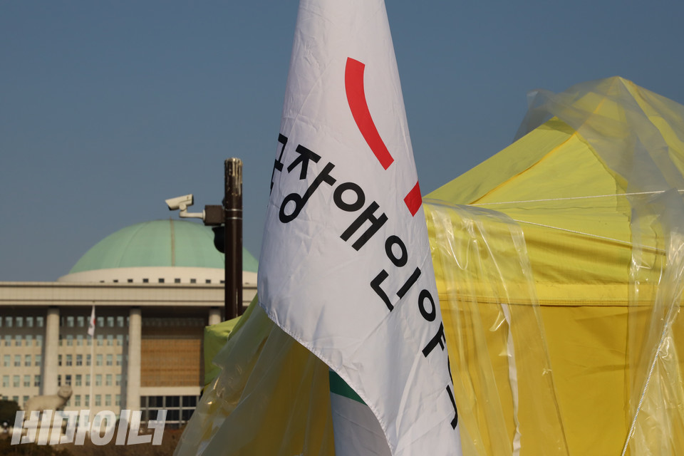 전국장애인야학협의회(전장야협)가 지난 16일 국회 앞에 설치한 노란색 농성장 천막과 깃발 뒤로 국회의사당 둥근 지붕이 보인다. 전장야협은 23일 오전 기자회견을 마친 뒤 일주일 만에 농성장을 정리했다. 사진 복건우