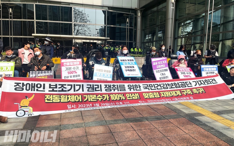 건보공단 서울강원본부 앞 기자회견 현장. 현수막에 “장애인 보조기기 권리 쟁취를 위한 국민건강보험공단 기자회견. 전동휠체어 기본수가 100% 인상! 맞춤형 지원체계 구축 촉구!”라고 적혀 있다. 사진 하민지