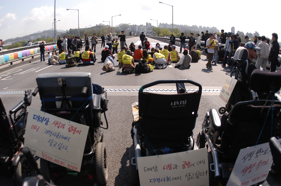 2006년 4월 27일, 중증장애인들이 활동보조제도화를 요구하며 한강대교를 6시간 동안 기었다. 텅 빈 휠체어가 보인다. 휠체어에는 “더 이상 시설과 골방이 아닌 지역사회에서 살고 싶다”는 피켓이 걸려 있다. 사진 김유미