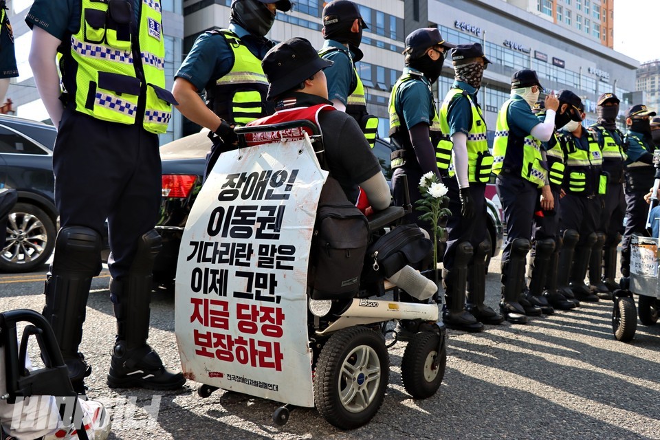 한 휠체어 이용 활동가가 농성역에서 하차해 행진을 준비하고 있다. 줄지어 선 경찰 옆에 있는 그는 휠체어 손잡이에 “장애인 이동권 기다리란 말은 이제 그만, 지금 당장 보장하라”라고 적힌 피켓을 걸었다. 사진 하민지