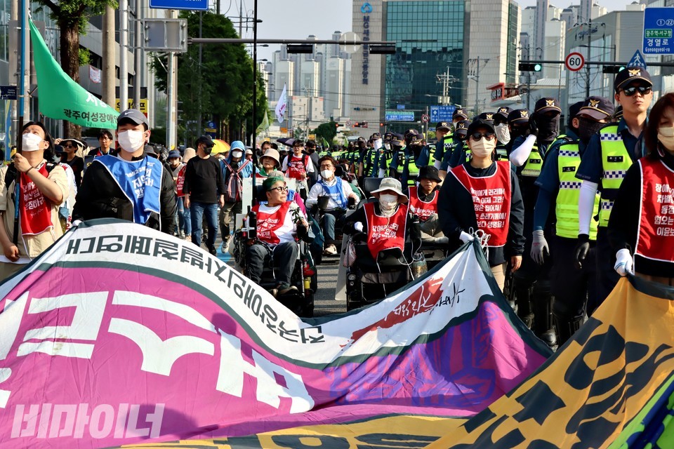 행진하는 활동가들. 대형 현수막 너머로 휠체어 이용 활동가들이 보인다. 제일 오른쪽에는 경찰이 줄지어서 걷고 있다. 사진 하민지
