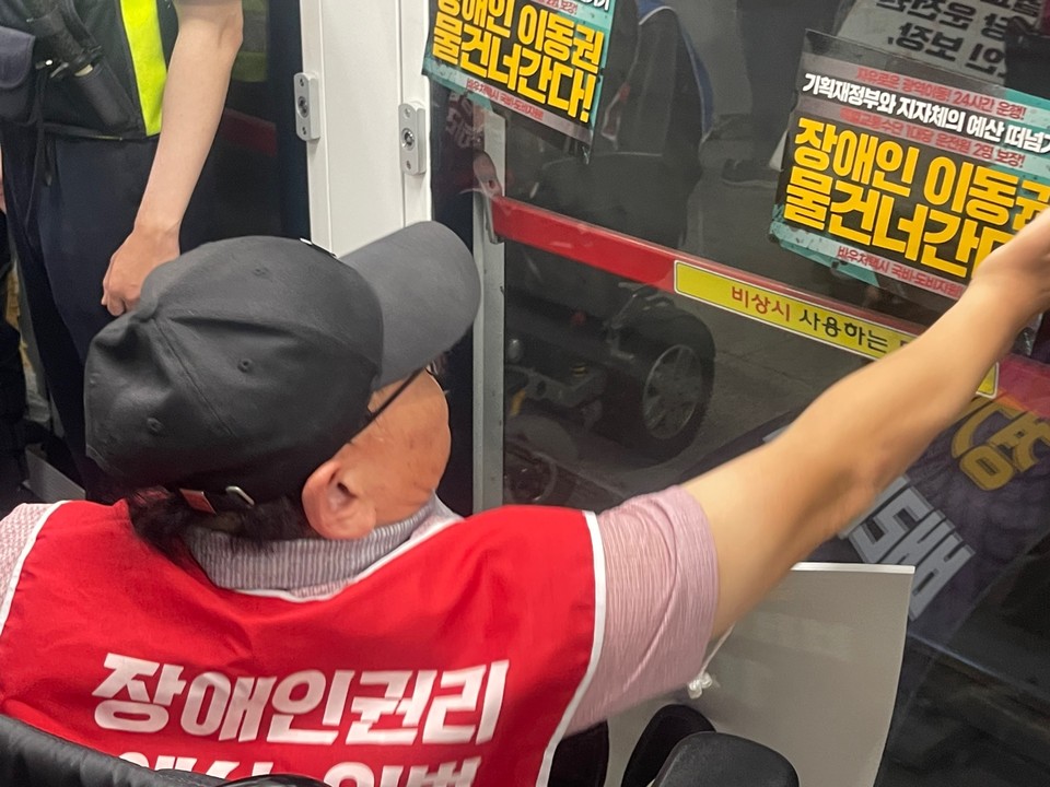 한 휠체어 이용 활동가가 안전문에 “기획재정부와 지자체의 예산 떠넘기기, 장애인 이동권 물 건너간다”라고 적힌 권리스티커를 붙이고 있다. 사진 경기장차연