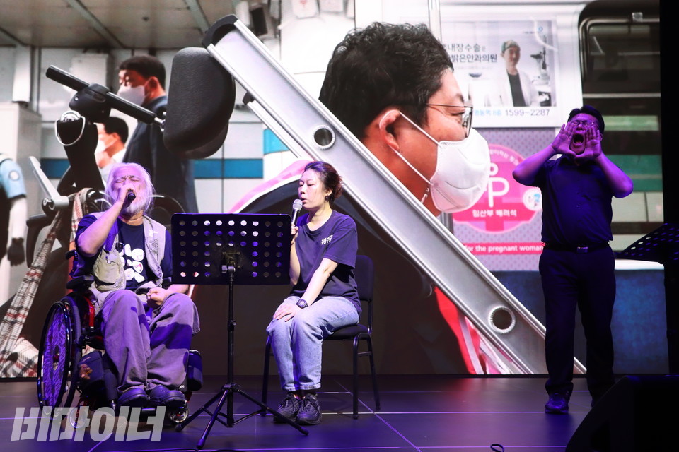 노들노래공장 수업을 하는 만수(가수 이민휘) 교사와 어깨꿈(박경석 전국장애인차별철폐연대 대표)이 ‘T4’ 노래를 부르고 있다. 그 뒤로 지하철 투쟁 영상이 펼쳐지고 있다. 사진 강혜민