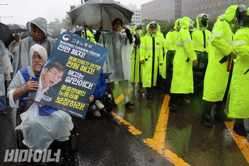 박경석 전장연 상임공동대표가 “고용노동부, 중증장애인 지역맞춤형 취업지원 전편 폐지! 동료지원가 187명 전원 해고! 해고는 살인이다! 중증장애인 노동권을 보장하라!”라고 적힌 피켓을 들고 있다. 그의 왼쪽으로 형광 우비를 입은 경찰 수십 명이 지나가고 있다. 사진 하민지