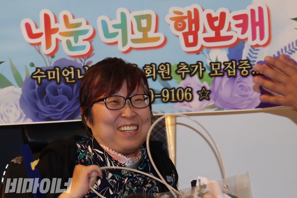 이수미 씨가 꽃다발을 받고 웃고 있다. 그의 뒤로 “나눈 너모 햄보캐(나는 너무 행복해)”라고 적힌 현수막이 보인다. 사진 하민지