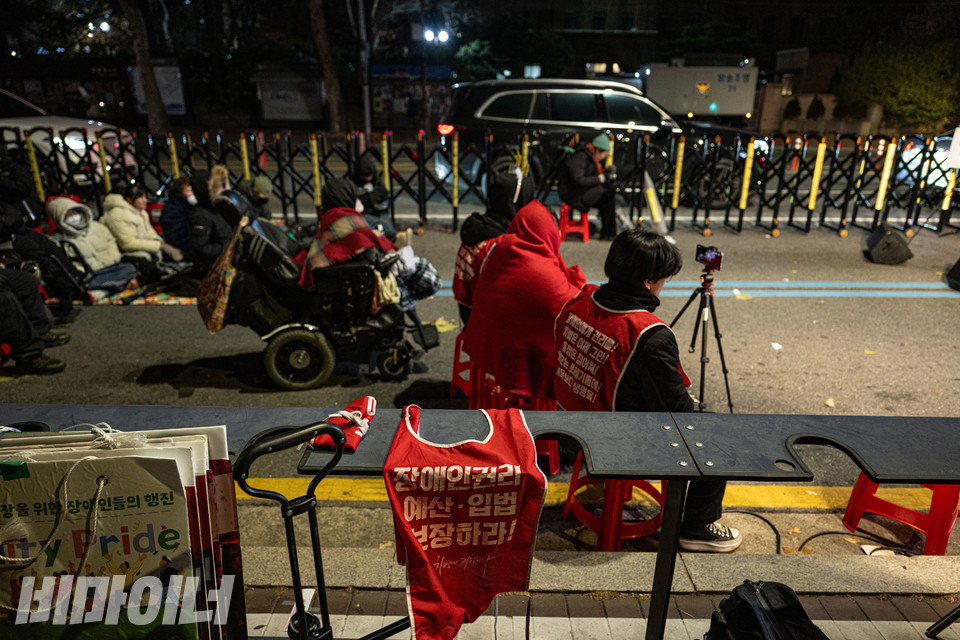 영하의 날씨 속에 담요로 몸을 꽁꽁 싸맨 활동가들이 보인다. 그 옆에 누군가 벗어 놓은 몸피씨가 있다. 몸피씨에는 ‘장애인권리예산·입법 보장하라!’고 적혀 있다. 사진 조진섭