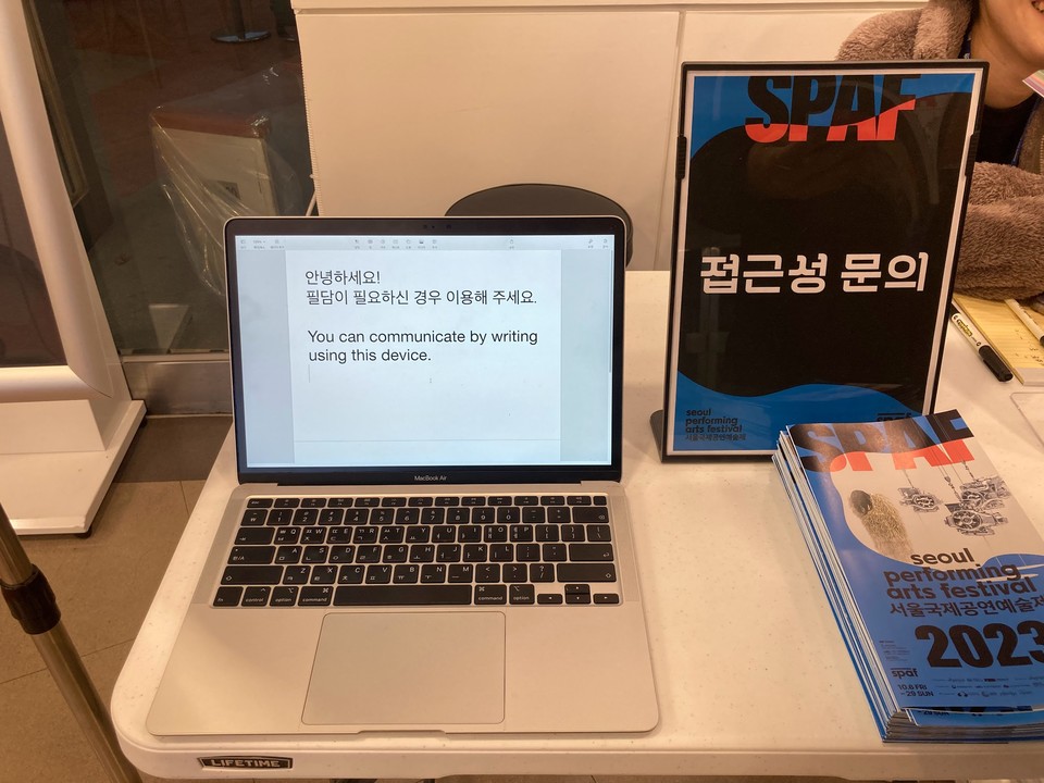 2023 SPAF 공연장의 접근성 데스크: 사진 중앙에 접이식 테이블이 놓여있다. 테이블 위에는 필담을 안내하는 문구가 크게 적힌 노트북이 한 대 있다. 그 오른쪽에는 SPAF 접근성 문의라고 적힌 알림 패널이 있고 뒤편에 앉아있는 스태프가 약간 보인다. ©2023 SPAF
