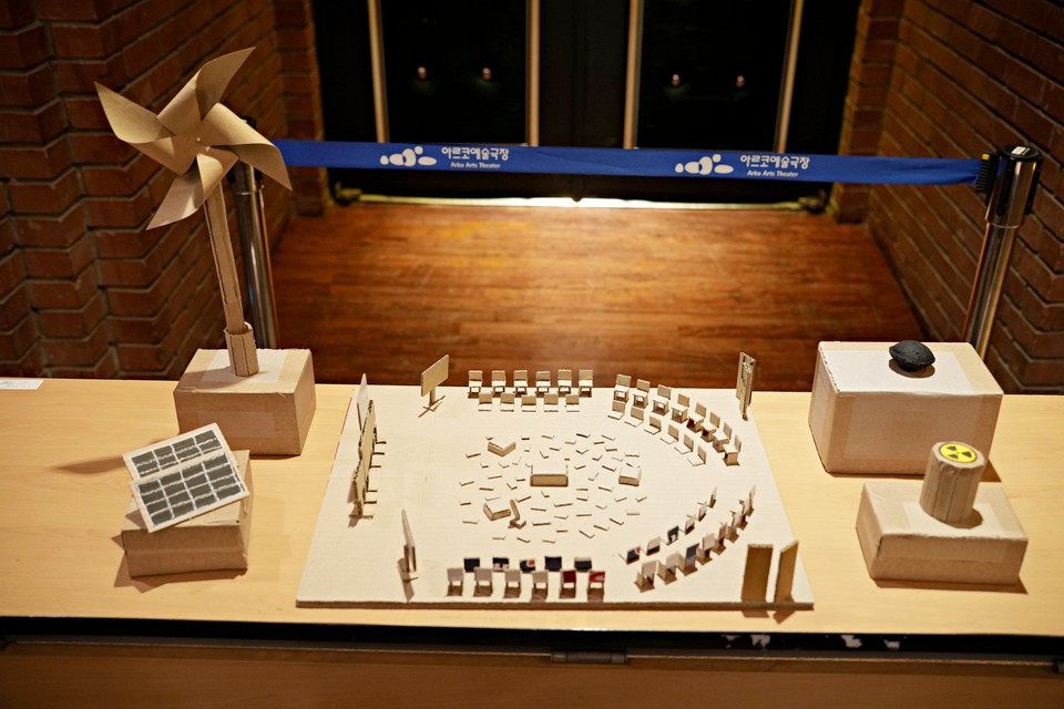 〈에너지_보이지 않는 언어〉 무대 축소 모형: 무대처럼 보이는 공간을 종이 소재로 재현한 모형이 전시되어 있다. 모형에는 두 줄의 의자들이 U자 형태로 둥글게 배치되어있다. 한 줄에 서른 석 정도다. 의자들 가운데 빈 공간에는 네모난 조각들과 상자 모양의 소품들이 수십 개 놓여있다. 맨 앞에는 네모난 칠판과 모니터 같은 것이 놓여있다. 무대 모형 좌측에는 바람개비와 태양광 전지 모양의 소품이, 우측에는 석유와 원자력을 나타내는 박스가 놓여있다. ©옥상훈·2023 SPAF