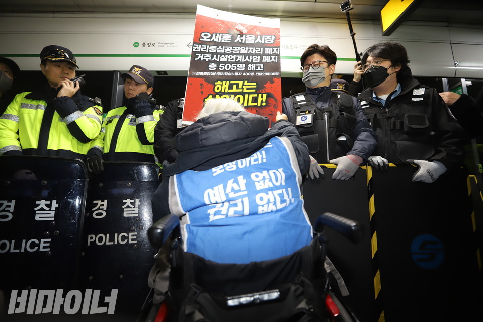박경석 대표가 “예산 없이 권리 없다”고 적힌 몸자보를 입고 있다. 경찰과 서울교통공사 직원이 그의 지하철 탑승을 가로막고 있다. 사진 강혜민