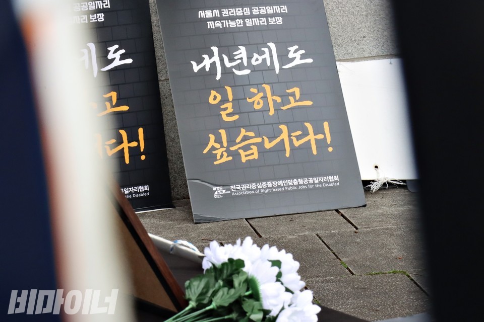 “서울시 권리중심공공일자리 지속가능한 일자리 보장, 내년에도 일하고 싶습니다”라고 적힌 피켓 아래로 국화꽃이 보인다. 사진 하민지