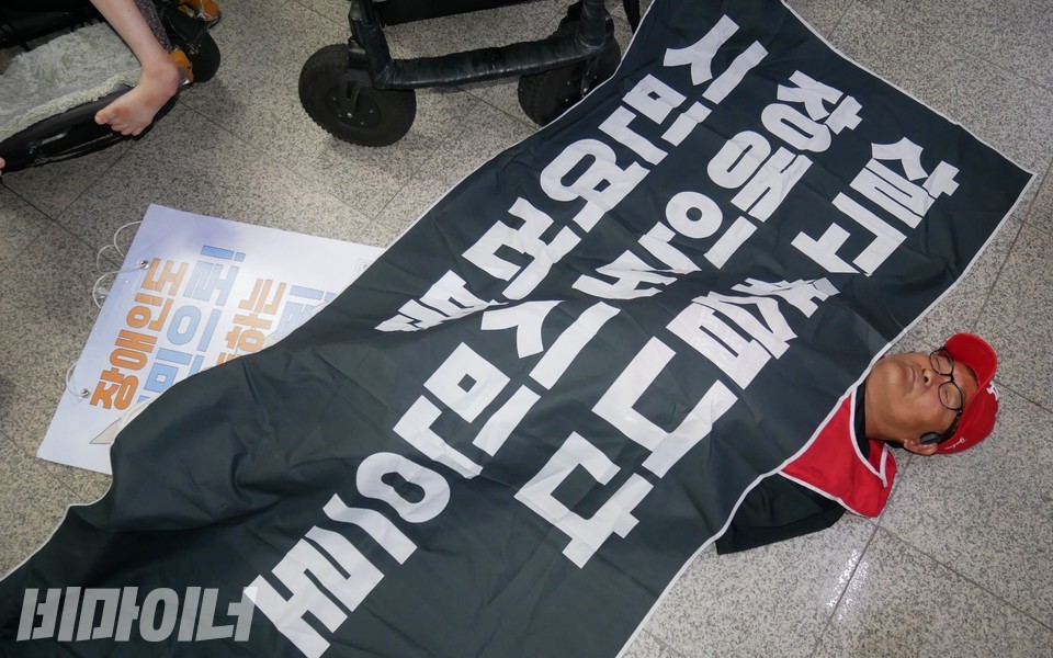 한 활동가가 국회의사당역 승강장 바닥에 누워 다이인 행동을 하고 있다. 그는 “시민 여러분 장애인도 시민으로 살고 싶습니다”라고 적힌 현수막을 덮고 있다. 사진 하민지