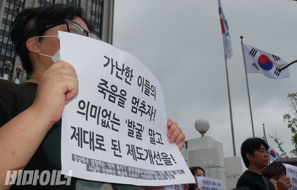 리버 홈리스야학 공동학생회장이 “가난한 이들의 죽음을 멈추자! 의미없는 발굴 말고 제대로 된 제도개선을!”이라 적힌 피켓을 들고 있다. 리버의 왼쪽에서 태극기가 휘날리고 있다. 사진 하민지