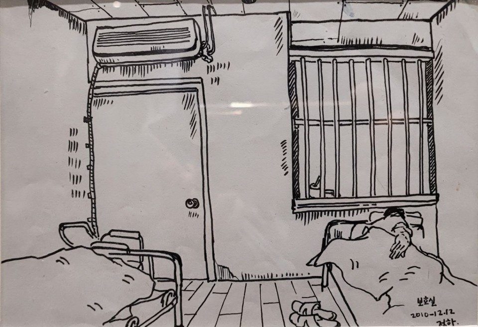보호실에 침대가 두 개 놓여 있다. 정면에 있는 벽 왼쪽에는 문이 있고, 오른쪽에는 쇠창살이 달린 창문이 있다. 그림 이정하(2010년)