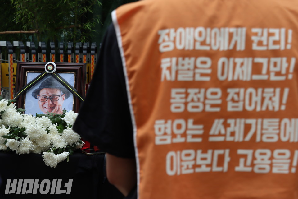 한 활동가가 영정을 바라 보고 있다. 그가 입은 주황색 몸 피씨에는 장애인 권리 보장을 촉구하는 문구가 쓰여 있다. 사진 강혜민