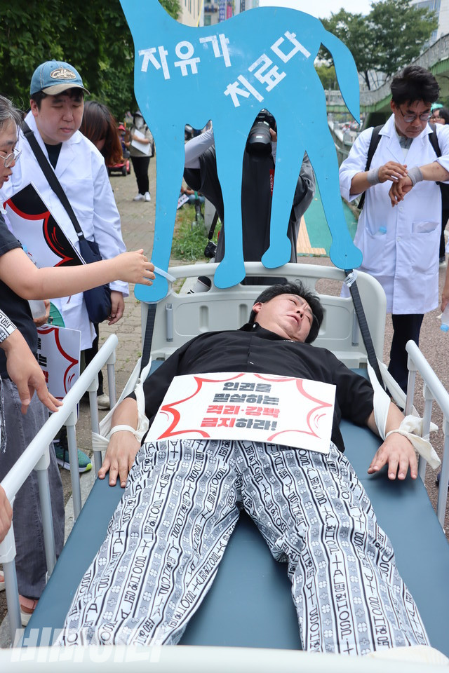 행진에 앞서 신석철 한국정신장애인연합회 상임대표가 침대에 손, 발이 묶여있는 퍼포먼스를 진행하고 있다. 그의 뒤에는 “자유가 치료다”라고 적힌 피켓이 있다. 사진 김소영