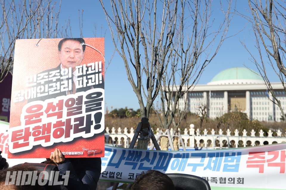 한 기자회견 참가자가 “민주주의 파괴! 장애인권리 파괴! 윤석열을 탄핵하라!”라고 적힌 피켓을 들고 있다. 그의 뒤로는 국회의사당 본관이 보인다. 사진 김소영
