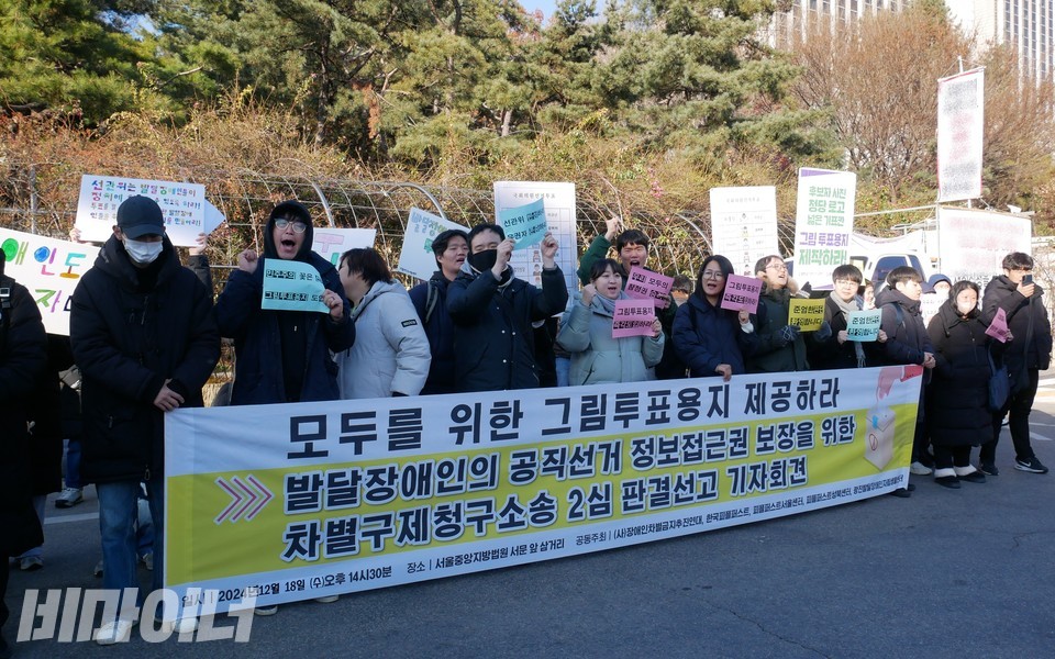 기자회견 현장. 참가자들이 함성을 지르고 있다. 현수막에는 “모두를 위한 그림투표용지 제공하라. 발달장애인의 공직선거 정보접근권 보장을 위한 차별구제청구소송 2심판결 선고 기자회견”이라 적혀 있다. 사진 하민지