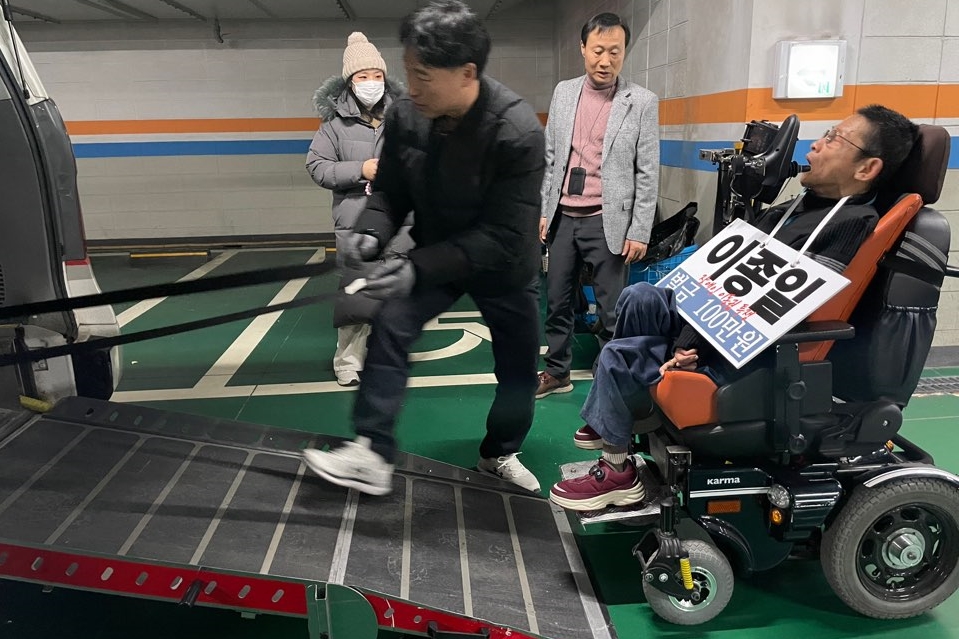 휠체어에 타고 있는 이종일 충북장차연 상임대표가 구치소로 이송되기 위해 차량에 탑승하고 있다. 이종일 대표의 목에는 자신의 이름과 “벌금 100만 원”이라고 적힌 피켓이 걸려있다. 사진 전장연 제공