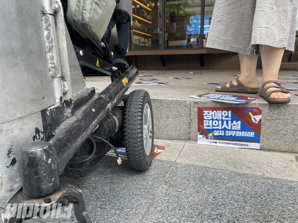 턱 왼쪽에는 턱을 오르지 못하고 있는 전동 휠체어의 바퀴가, 턱 위에는 비장애인의 발이 보인다. “장애인 편의시설 설치 의무화하라!”라고 적힌 스티커가 턱에 붙어 있다. 사진 하민지