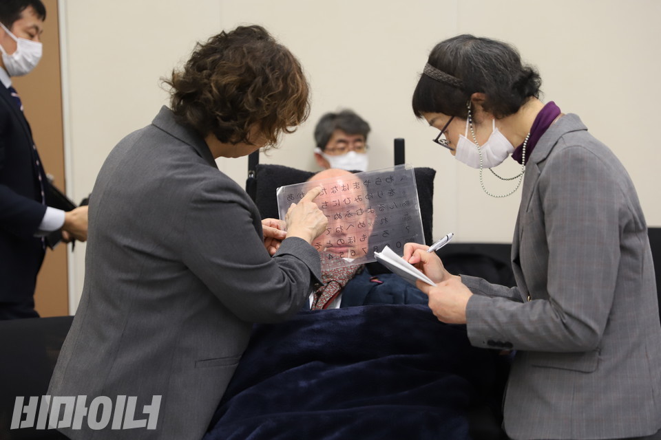 후나고 야스히코 의원이 눈동자를 움직여 지원인이 들고 있는 일본어가 적힌 판에서 글자를 선택하고 있다. 두 명의 지원인 중 한 지원인(왼쪽)은 일본어가 적힌 판에서 글자를 가리키고 있고, 다른 지원인(오른쪽)은 글자를 받아적고 있다. 사진 김소영