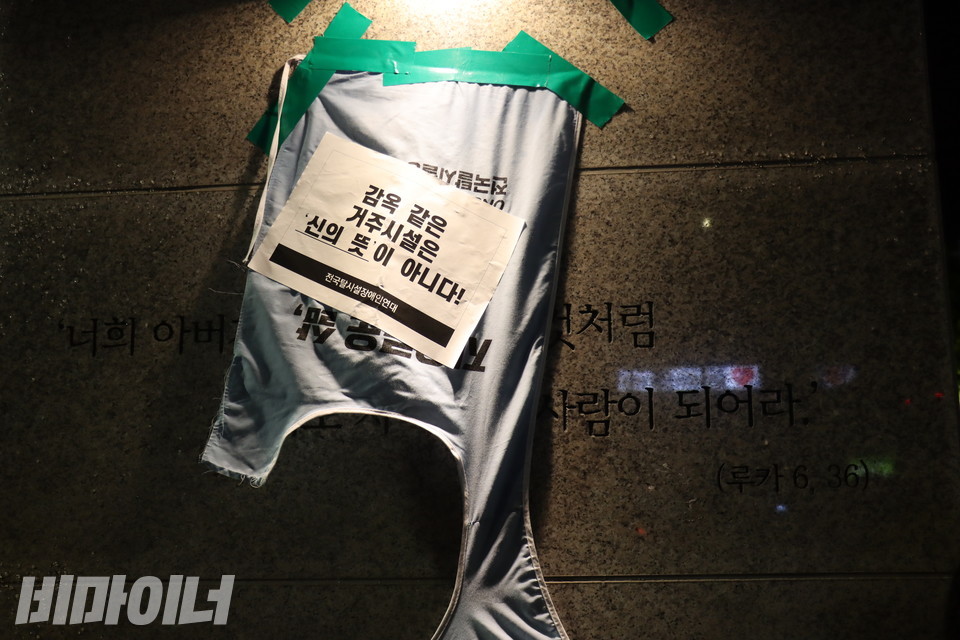 활동가가 성당 문 옆 기둥에 붙여둔 몸피씨를 성당 관계자가 거칠게 뜯어냈다. 이후 참여자들은 뜯긴 몸피씨를 청테이프로 다시 붙였다. 몸피씨 위에는 “감옥 같은 거주시설은 신의 뜻이 아니다!”라는 문구가 적힌 스티커가 붙어 있다. 이 몸피씨는 기둥에 새겨진 “너희 아버지께서 자비하신 것처럼 너희도 자비로운 사람이 되어라”라는 문장을 가리고 있다. 사진 김소영