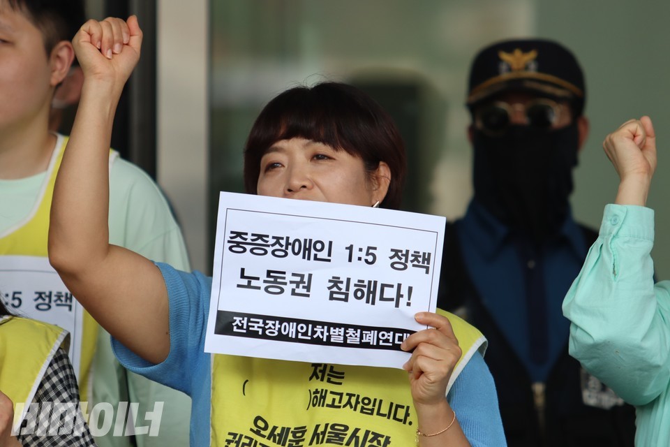 기자회견 참여자가 한 손으로는 “중증장애인 1:5 정책 노동권 침해다!”라고 적힌 종이 피켓을 들고, 다른 한 손은 높이 들며 구호를 외치고 있다. 사진 김소영 