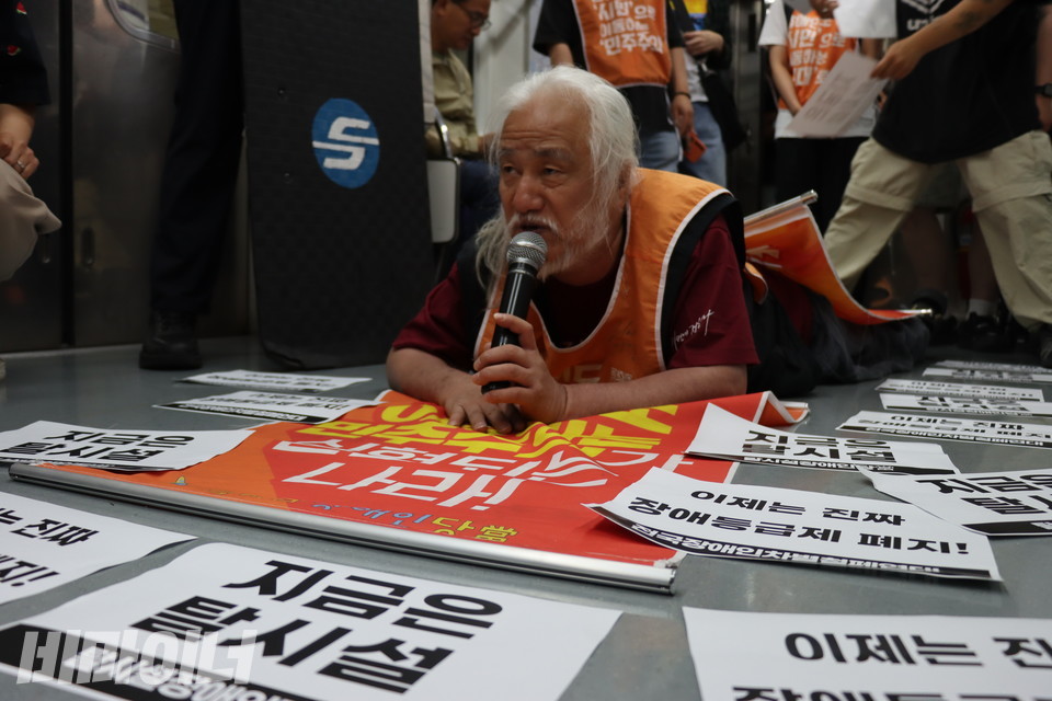 박경석 전장연 상임공동대표가 포체투지를 한 채 마이크를 쥐고 발언하고 있다. 그의 주변 바닥에는 “이제는 진짜 장애등급제 폐지!”, “지금은 탈시설” 등이 적힌 스티커가 붙어 있다. 사진 김소영