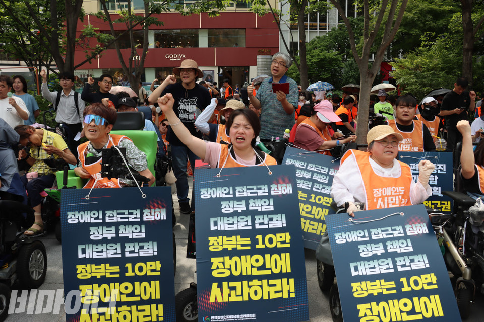 기자회견 참여자들이 손을 높이 들고 구호를 외치고 있다. 그들의 목에는 “장애인 접근권 침해 정부 책임 대법원판결! 정부는 10만 장애인에 사과하라!”라고 적힌 피켓이 걸려 있다. 사진 김소영