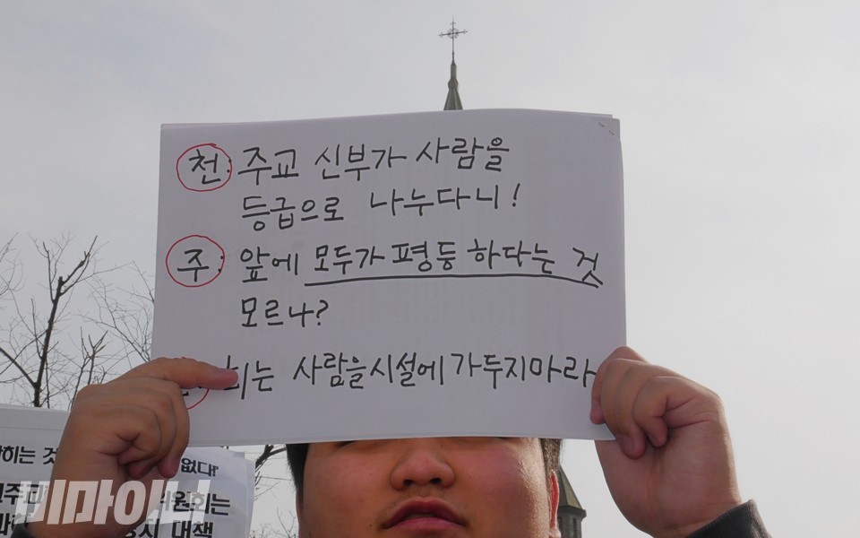 2023년 12월 10일, 한 활동가가 기자회견에서 든 피켓에 천주교 삼행시가 적혀 있다. ‘천’주교 신부가 사람을 등급으로 나누다니! ‘주’ 앞에 모두가 평등하다는 것 모르나? ‘교’회는 사람을 시설에 가두지 마라. 사진 하민지