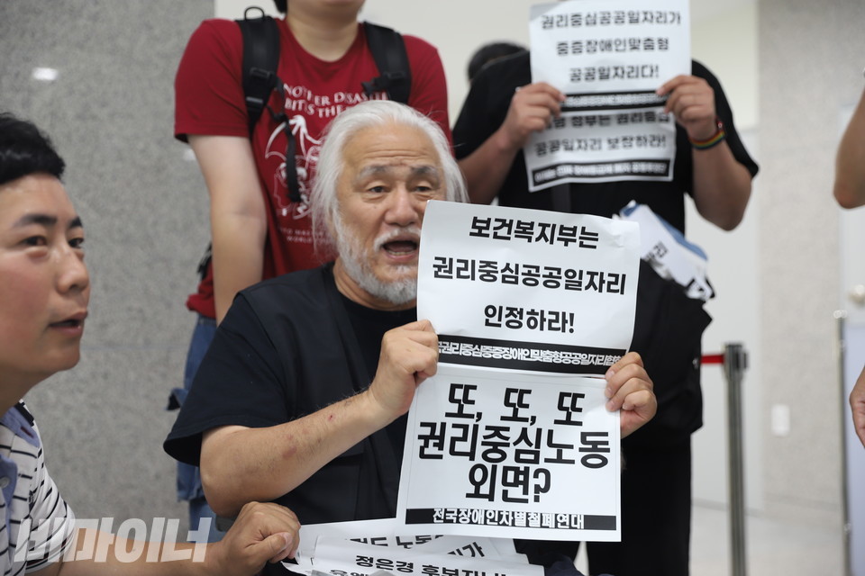 국정기획위원회에서 긴급행동을 벌이는 박경석 전장연 상임공동대표가 종이 피켓을 들고 있다. 종이에는  “보건복지부는 권리중심공공일자리 인정하라!”,  “또, 또, 또 권리중심노동 외면?”이라고 적혀있다. 사진 김소영
