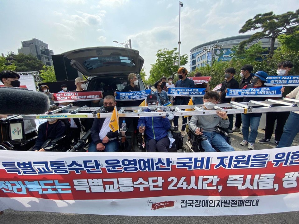 휠체어를 탄 장애인들이 장애인콜택시를 막아서고 사다리를 목에 걸고 있다. 사다리에는 "특별교통수단 운영예산 국비 지원하라!"라는 현수막을 걸었다. 사진 전국장애인이동권연대