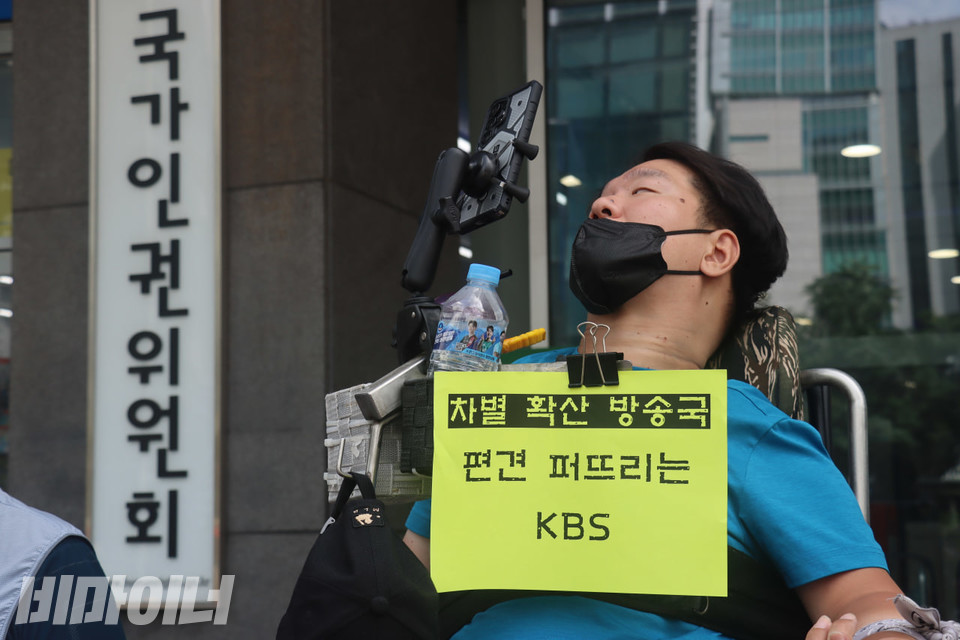 전동휠체어를 이용하는 장애인 활동가가 기자회견에 참석한 모습. 그의 휠체어에 “차별 확산 방송국. 편견 퍼뜨리는 KBS”라고 적힌 형광색 종이 피켓이 붙어 있고, 그의 뒤로는 ‘국가인권위원회’ 간판이 보인다. 사진 김소영