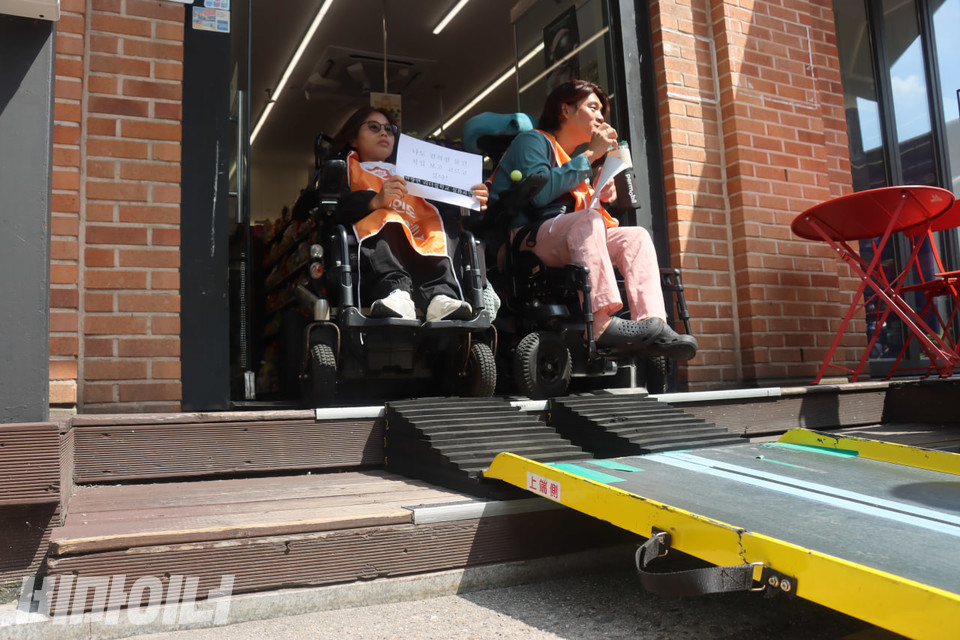 CU편의점 마로니에공원점 입구. 계단에 스티로폼 경사로와 이동식 경사로가 놓여 있다. 계단 위에는 두 명의 휠체어 이용 장애인 활동가들이 있다. 사진 김소영