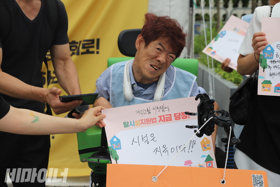 김탄진 탈시설연대 서울지부 활동가가 AAC(보완대체 의사소통기기)를 통해 발언하고 있다. 그의 앞에는 “시설은 지옥이다!!”라고 적힌 종이 피켓이 놓여 있다. 사진 김소영