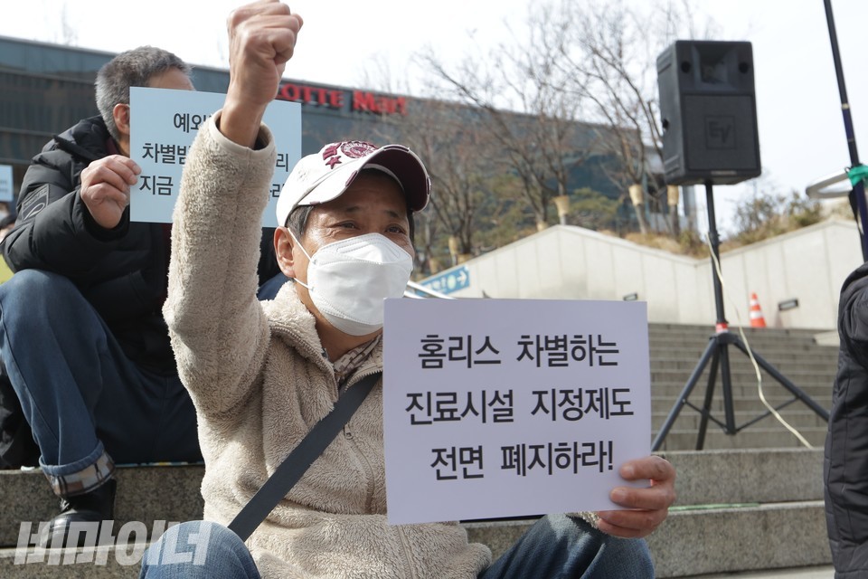 2022년 3월 10일, 서울역 광장에서 열린 결의대회에서 자물쇠 씨가 ‘홈리스 차별하는 진료시설 지정제도 전면 폐지하라’라고 적힌 피켓을 들고 ‘투쟁’을 외치고 있다. 사진 비마이너