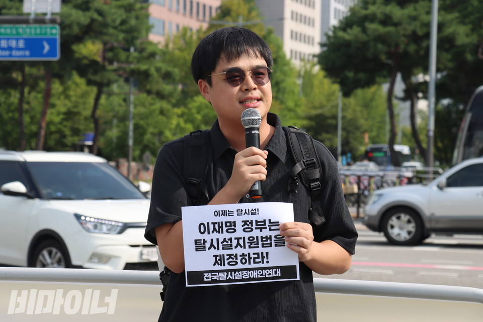 국회 앞에서 열린 기자회견에서 이한결 한국정신장애인연합회 활동가가 발언하고 있다. 그의 손에는 “이제는 탈시설! 이재명 정부는 탈시설지원법을 제정하라!”라고 적힌 종이 피켓이 들려 있다. 사진 김소영