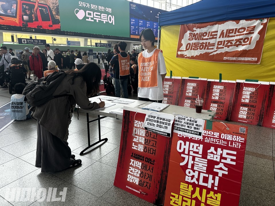 활동가들이 천막 농성장 앞에 책상을 펼치고, 교통약자이동권보장법 제정을 위한 시민 서명을 받고 있다. 한 시민이 서명을 하고 있다.