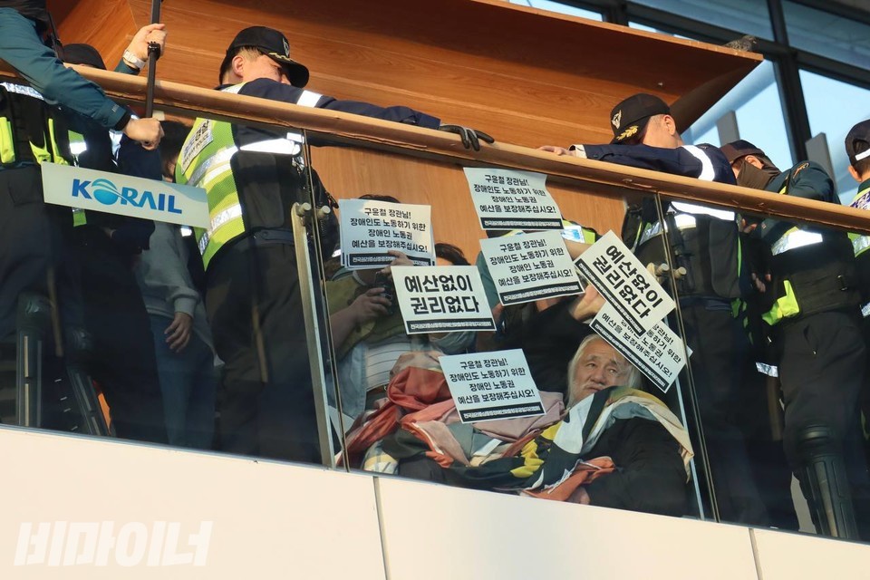 대합실 위층 난간에서 현수막을 내리기 위한 시도를 하던 박경석 전국장애인차별철폐연대 상임공동대표가 경찰에 둘러싸여 있다. 허망한 표정을 짓고 있는 박 대표의 앞에 “예산 없이 권리 없다”라고 적힌 스티커가 유리창에 붙어 있다. 사진 김소영