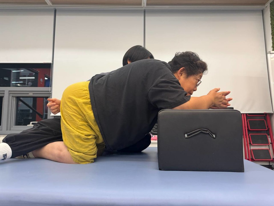 형숙이 사각 폼 블록을 팔꿈치 아래에 받치고 네발서기 자세를 하고 있다. 직각으로 굽힌 형숙의 다리가 들리지 않도록 PT선생님이 누르고 있다. 사진 탁영희