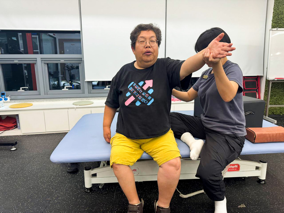 형숙이 침대에 걸터앉아 왼팔을 쭉 펴서 앞으로 뻗고 있다. PT선생님은 형숙의 중심이 흐트러지지 않도록 팔과 허리를 받쳐준다. 사진 탁영희