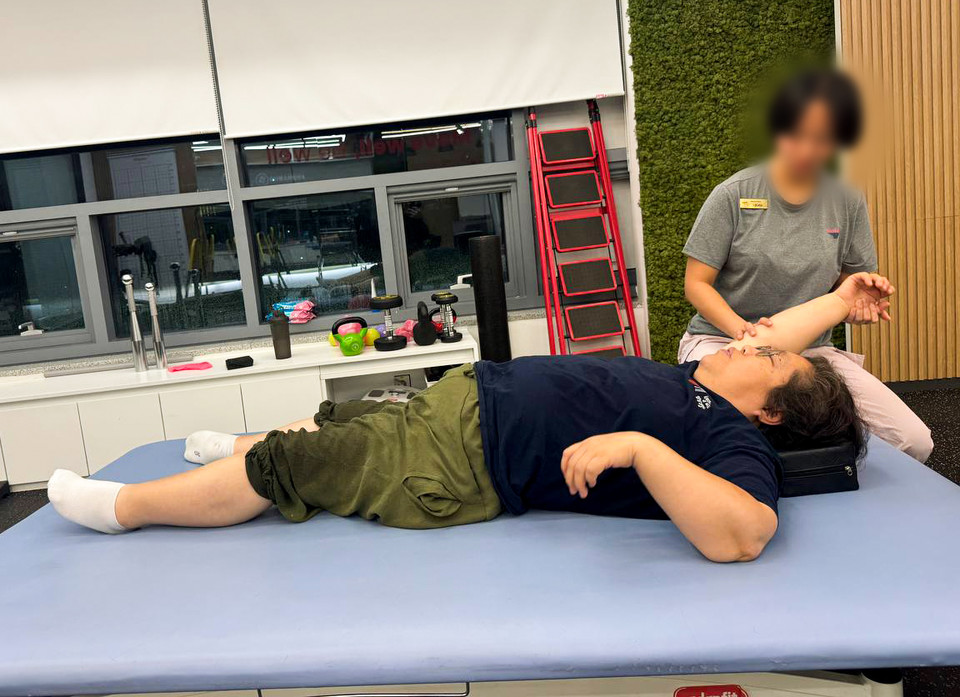 형숙이 침대에 누워 오른팔을 귀 옆쪽으로 들어 올리고, PT선생님이 형숙의 팔이 떨어지지 않도록 받쳐주고 있다. 형숙의 바지 밑단은 젖어있다. 사진 탁영희