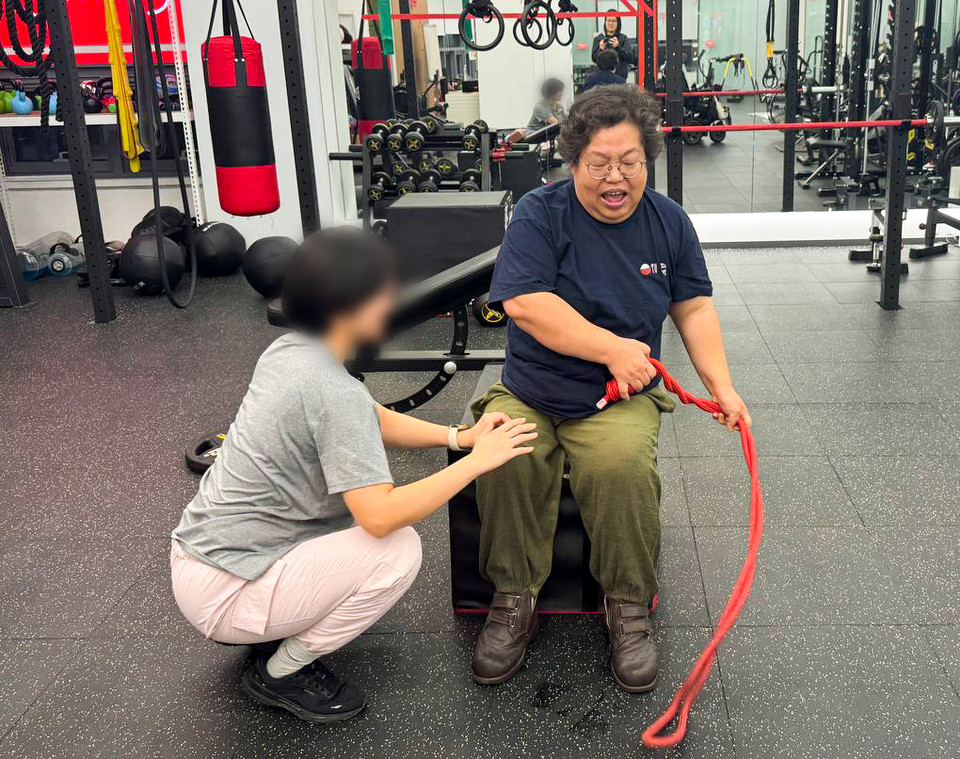 형숙이 빨간 줄을 두 손으로 잡고 돌리고 있다. PT선생님이 형숙의 오른쪽 다리가 들리지 않도록 붙잡고 있다. 사진 탁영희