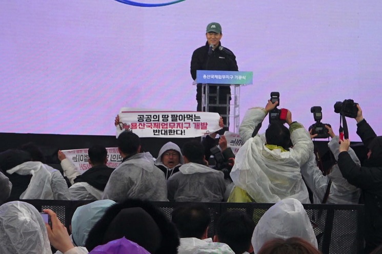 오세훈 시장이 기공식에서 비전 선포를 하는 순간 기습적으로 항의 행동에 나선 시민들. 한 활동가가 “공공의 땅 팔아먹는 용산국제업무지구 개발 반대한다!”라고 적힌 손 피켓을 높이 들고 있다. 그 뒤로 웃고 있는 오세훈 시장의 모습이 보인다. 사진 용산정비창 개발의 공공성 강화를 위한 공동대책위원회