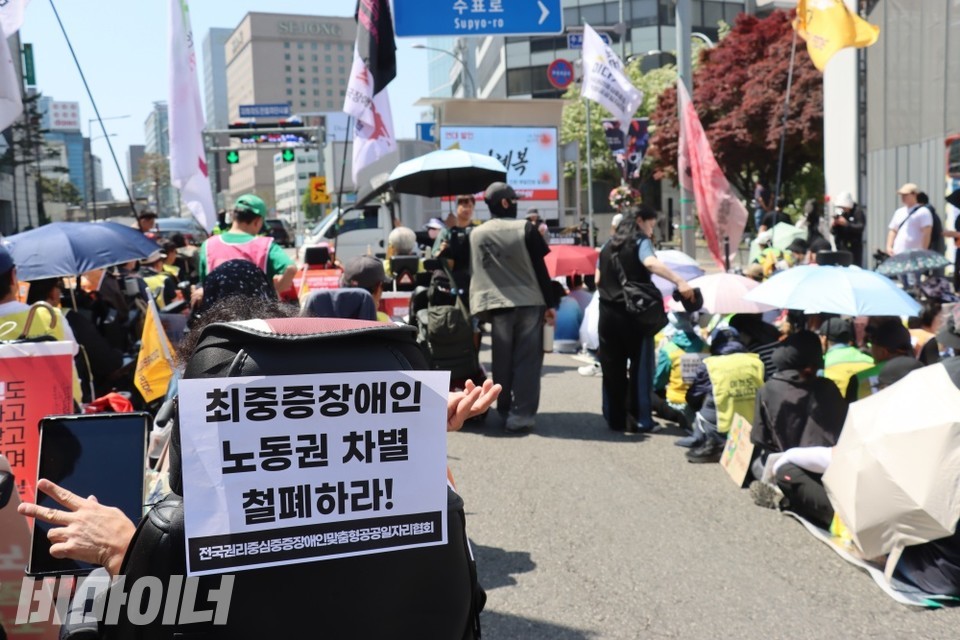 장애인노동절 결의대회 현장. 휠체어에 "최중증장애인 노동권 차별 철폐하라!" 스티커가 붙여져 있고, 그 너머로 무대차량이 보인다. 사진 이재민