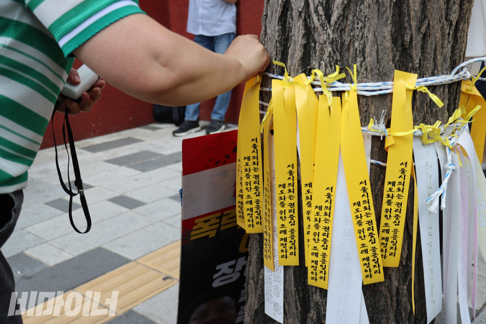 한 결의대회 참가자가 “서울시의회는 권리중심공공일자리 제도화를 위한 조례 제정하라”라고 적힌 노란끈을 나무 위의 밧줄에 묶고 있다. 사진 김소영
