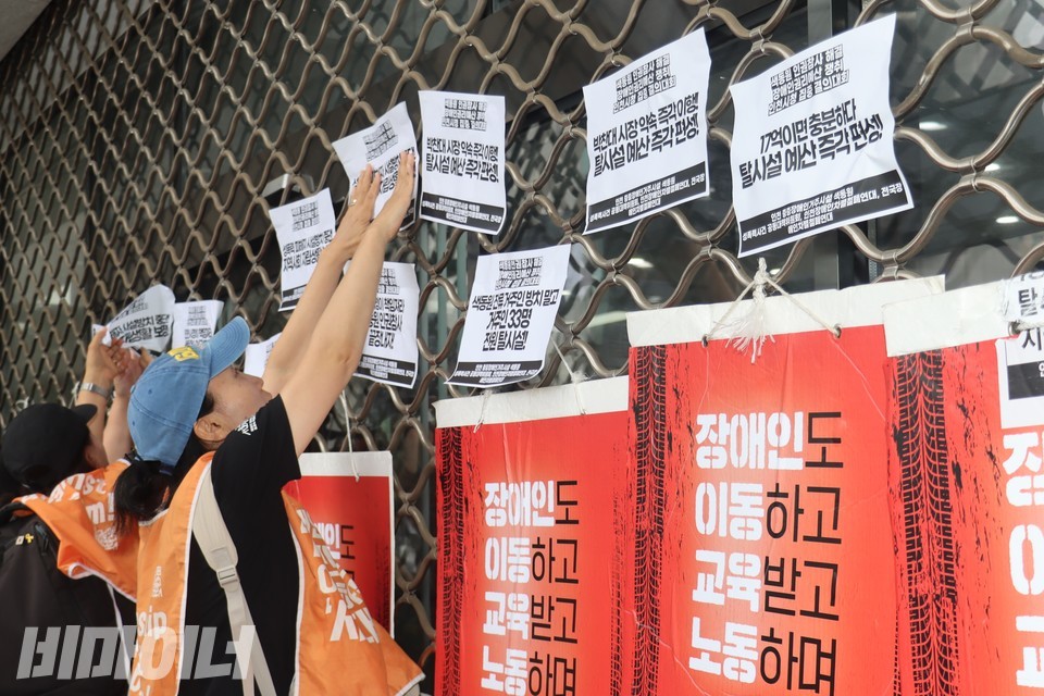 결의대회 참가자가 정문에 내려온 철제 셔터에 공대위 요구가 담긴 스티커를 붙이고 있다. 스티커에는 “박찬대 시장 약속 즉각 이행! 탈시설 예산 즉각 편성”, “17억이면 충분하다” 등의 문구가 쓰여있다. 사진 김소영